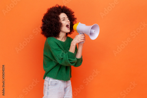 Side view portrait of woman with Afro hairstyle wearing green casual style sweater protesting or making announcement, screaming in megaphone. Indoor studio shot isolated on orange background.