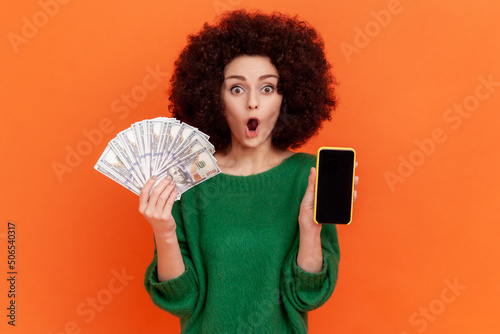 Portrait of shocked woman with Afro hairstyle wearing green casual sweater holding fan of hundred dollars bills and cell phone with empty display. Indoor studio shot isolated on orange background.