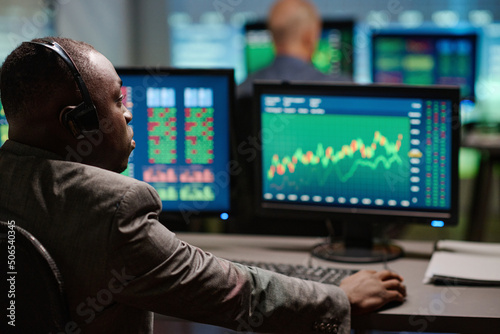 Rear view of African American man sitting in front of computer monitors watching and analyzing trading stats in evening