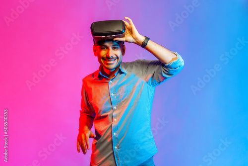 Portrait of smiling man in shirt wearing virtual reality goggles  raising VR headset and screaming with positive expression. Indoor studio shot isolated on colorful neon light background.