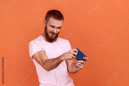 Portrait of handsome young adult bearded man standing, using smartphone and playing mobile game with frowning face, wearing pink T-shirt. Indoor studio shot isolated on orange background. © khosrork