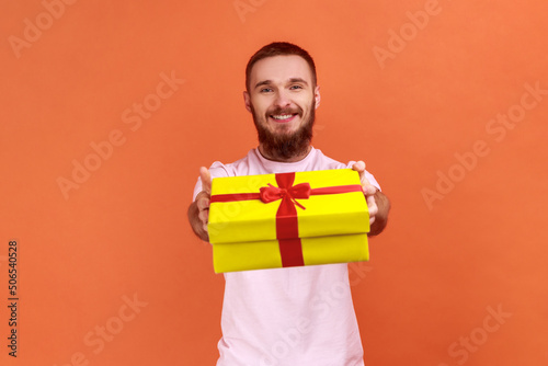 Portrait of happy bearded man giving gift box to camera with excited smile, greeting on holiday and sharing present, wearing pink T-shirt. Indoor studio shot isolated on orange background. © khosrork