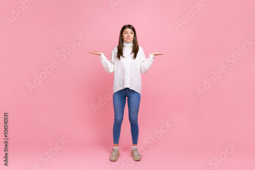 Full length portrait of helpless pretty female with spread arms, does not know answer on questions, wearing white casual style sweater. Indoor studio shot isolated on pink background. photo