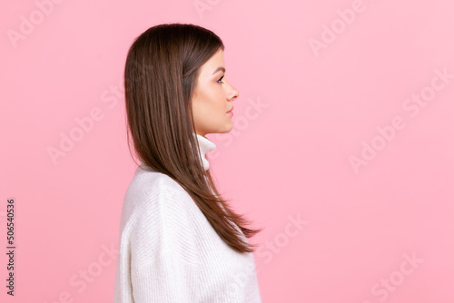 Side view portrait of serious female looks ahead with strict expression, expressing negative emotions, wearing white casual style sweater. Indoor studio shot isolated on pink background.