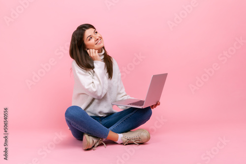 Portrait of female with dreamy facial expression, sitting with crossed legs on floor, holding laptop, wearing white casual style sweater. Indoor studio shot isolated on pink background.