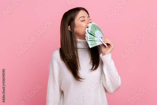 Tableau sur toile Positive greedy girl with dark hair holding big fan of euro banknotes, smelling money, lottery winner, wearing white casual style sweater