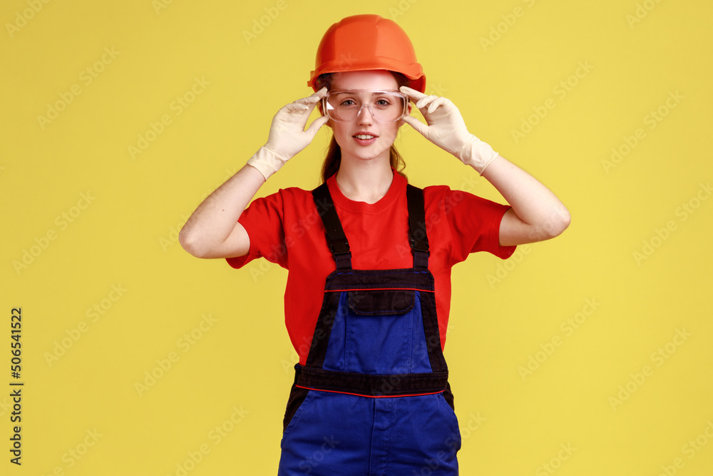 Portrait of confident builder woman standing putting on protective glasses, looking at camera, wearing overalls and protective helmet. Indoor studio shot isolated on yellow background.