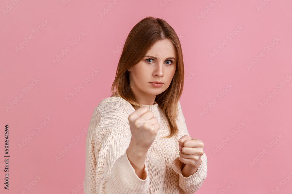 Portrait of angry confident attractive blond woman clenching fists, showing boxing gesture and ready to punch, wearing white sweater. Indoor studio shot isolated on pink background. Portrait of angry confident attractive blond woman clenching fists, showing boxing gesture and ready to punch, wearing white sweater. Indoor studio shot isolated on pink background.