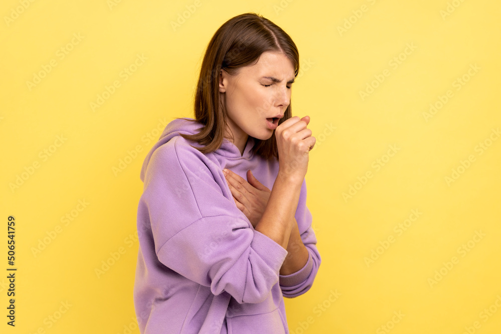 Portrait of young adult sick woman with dark hair have cough and feeling pain in her lungs, symptoms of disease, wearing purple hoodie. Indoor studio shot isolated on yellow background. Portrait of young adult sick woman with dark hair have cough and feeling pain in her lungs, symptoms of disease, wearing purple hoodie. Indoor studio shot isolated on yellow background.