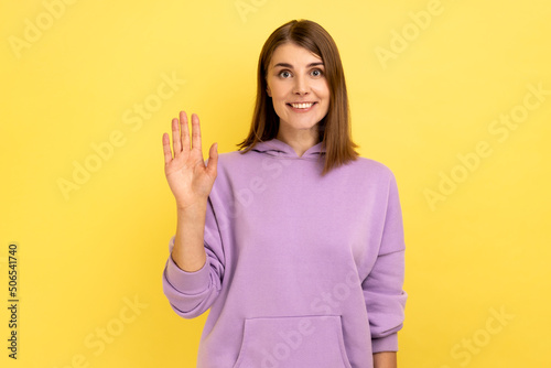 Portrait of friendly positive young woman smiles toothily  raises palm greets friend  saying hello or goodbye  wearing purple hoodie. Indoor studio shot isolated on yellow background.
