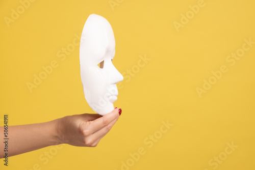 Profile side view closeup of woman hand holding white facial mask model, hiding personality. Indoor studio shot isolated on yellow background. photo