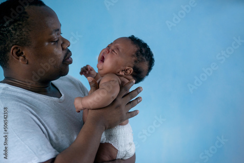 African father holding and comforting sad baby daughter against blue background, holding crying little newborn baby in his arms photo