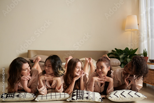 Positive young women in satin pajamas lying on floor and smiling at each other when enjoying sleepover