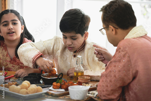 Indian father and daughter and son eating Indian food, using hand, panipuri, curry butter chicken, chicken Tikka, biryani, and naan bread on wooden table, Indian family photo