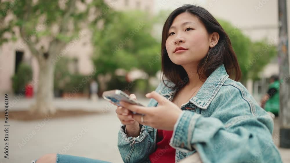 Long haired Asian woman wearing denim jacket sitting on the bench outdoors checking her smartphone and looking for friend that must come Long haired Asian woman wearing denim jacket sitting on the bench outdoors checking her smartphone and looking for friend that must come