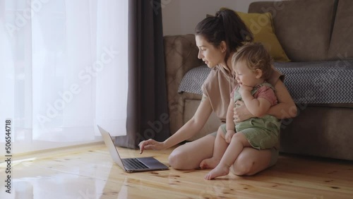 Mother multi-tasking, holding baby infant and using computer laptop at home. Candid authentic and real life mom working and parenting. young little toddler boy and baby playing together around mother