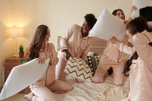 Joyful young women in silk pajamas fighting with pillows on bed photo