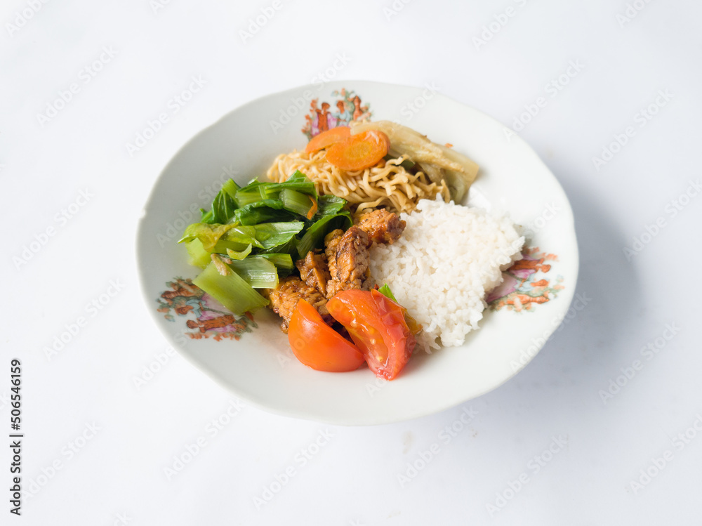 Cooked white rice, fried noodles on a white plate, topped with sliced green vegetables plus tomatoes and sliced fried tempeh