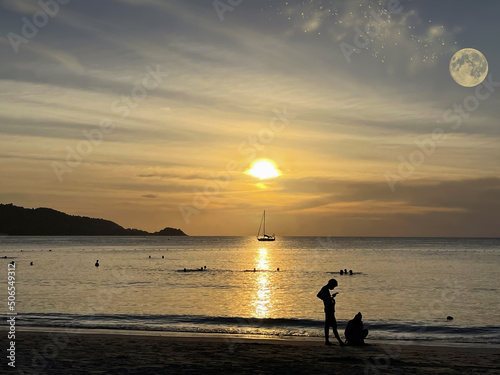 Sunset over the sea. People on the beach. Swimming people. Beautiful sky with clouds  sun  stars  moon. Reflection on the water surface. Golden glow  sunshine. Sand coast. Tropical paradise. Island.  