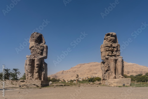 Giant statues of the colossi of Memnon against the blue sky and sand dunes. Huge damaged statues of pharaohs sitting on pedestals. Egypt