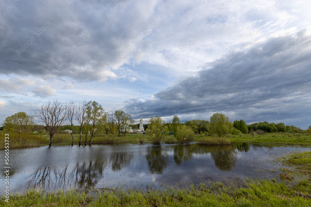 Rural landscape on a spring evening. Dramatic sky reflected in the river. Juicy green grass in the foreground.