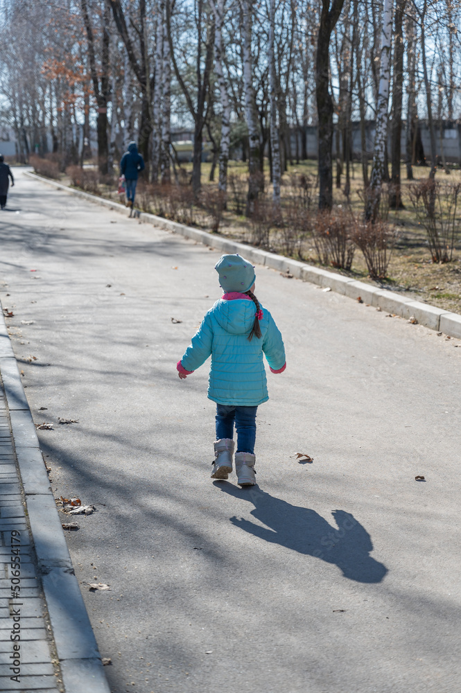 A child runs through a spring park. A five-year-old girl with long pigtails in a turquoise jacket. View from the back.