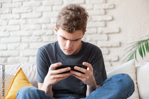 A teenage boy plays games on a smartphone with a focused expression. Dependence on the phone and online games. Close-up, bottom view. A young man with a phone in his hands sits on the couch.