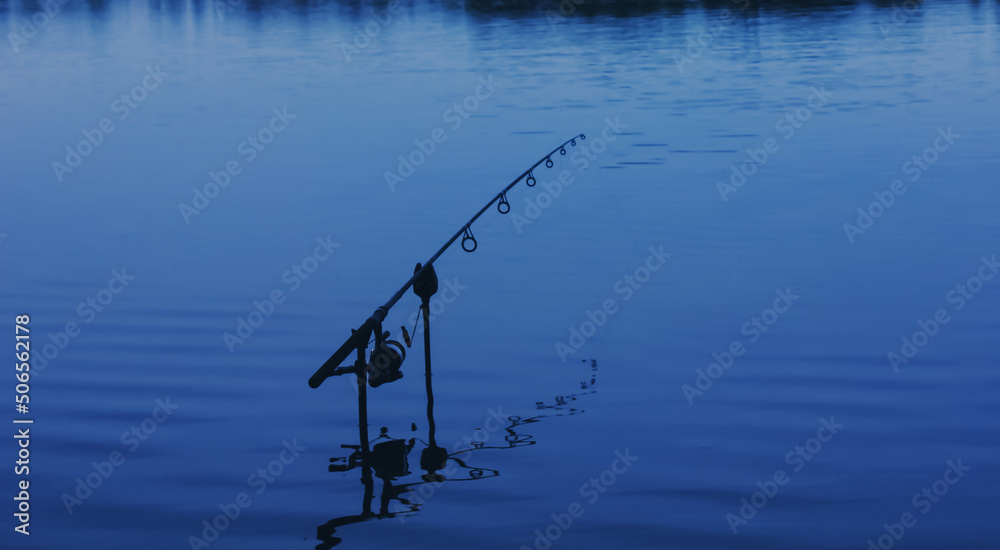 Single fishing rod mounted on banksticks with a bite alarm during a session of carp fishing. Fishing at dusk and into the night. Abstract defocused image.