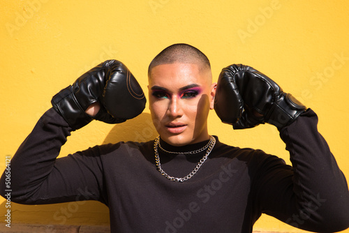 Tablou pe pânză Young non-binary person wears boxing gloves on a mustard yellow background, the person is make up and does different boxing poses