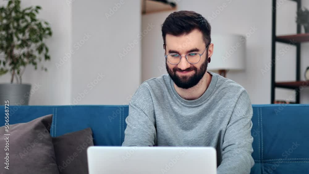 Smiling handsome Hispanic hipster man working chatting remotely use laptop sitting on blue couch Smiling handsome Hispanic hipster man working chatting remotely use laptop sitting on blue couch