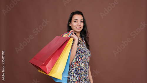 Young adult holding colorful shopping bags on camera in studio, after buying clothes on discount sale. Shopaholic female model making purchase at retail store to buy things in paper bags.