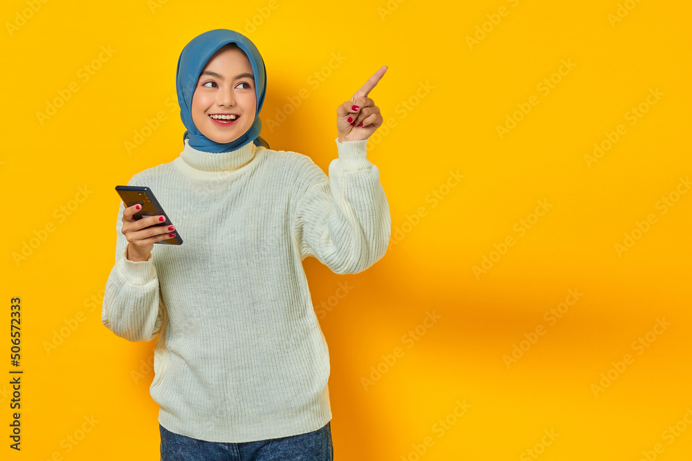 Portrait of smiling young asian woman in white sweater and hijab holding mobile phone, pointing finger aside isolated over yellow background. People islam religious concept Portrait of smiling young asian woman in white sweater and hijab holding mobile phone, pointing finger aside isolated over yellow background. People islam religious concept