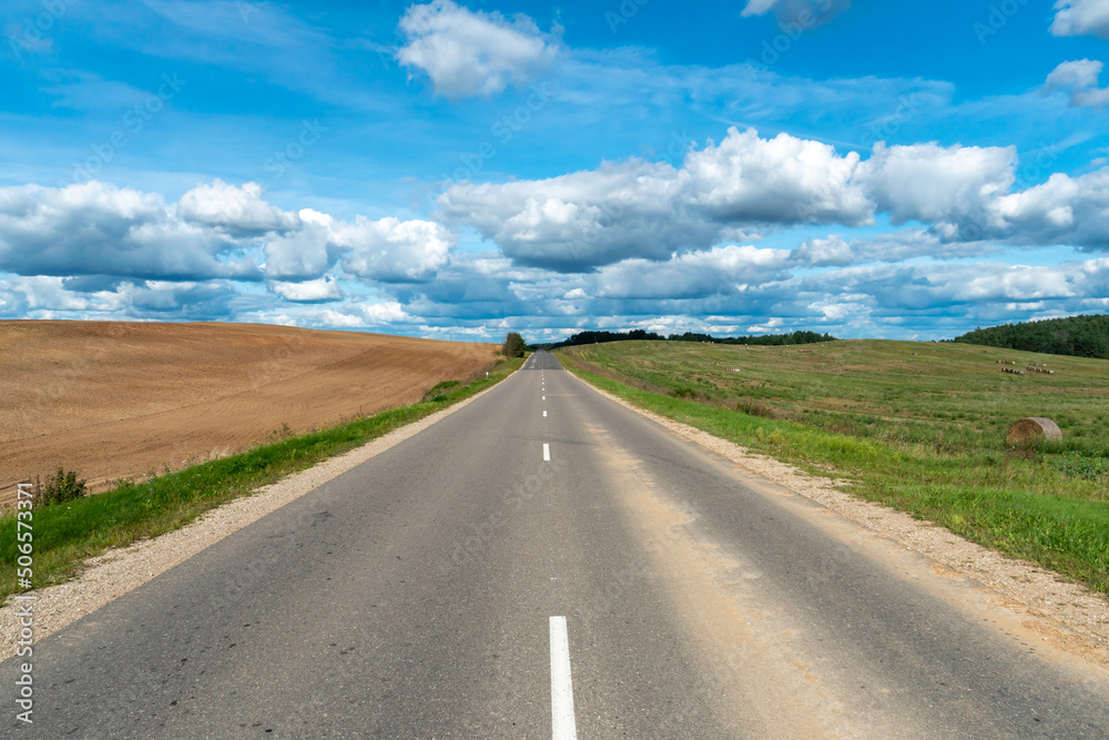 An asphalt road section in a rural area passing by agricultural fields against a background of blue sky and white fluffy clouds on a warm sunny day.