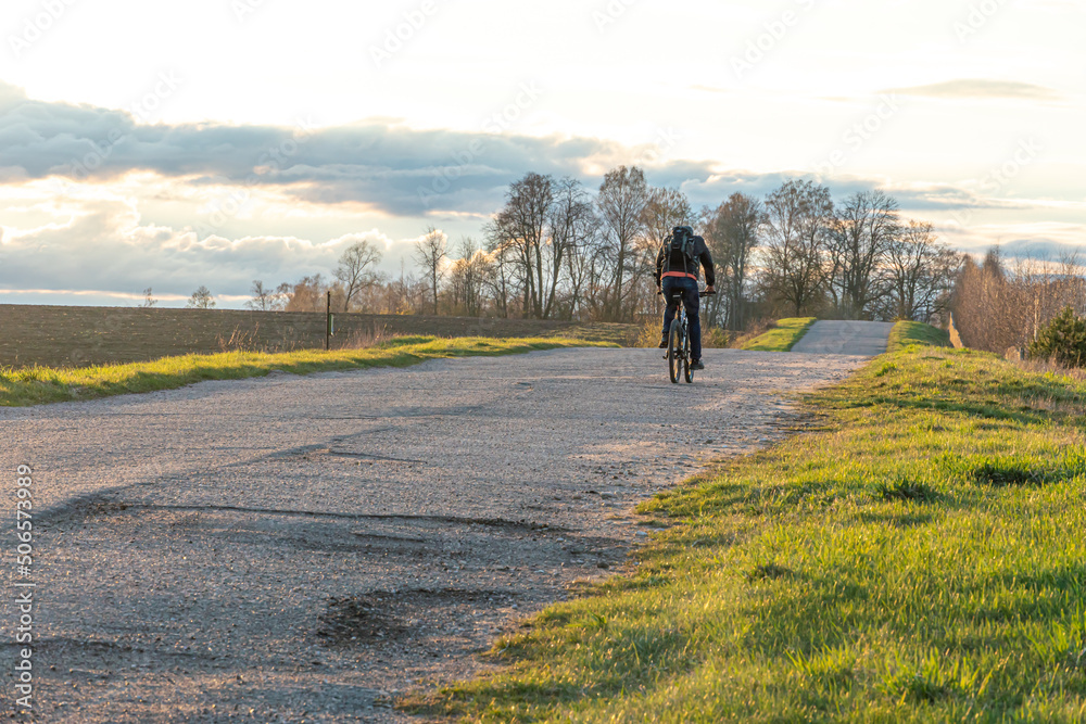 Obraz premium A cyclist rides on an asphalt road in the countryside during sunset past beautiful fields and flower meadows. A receding single cyclist on an empty road.