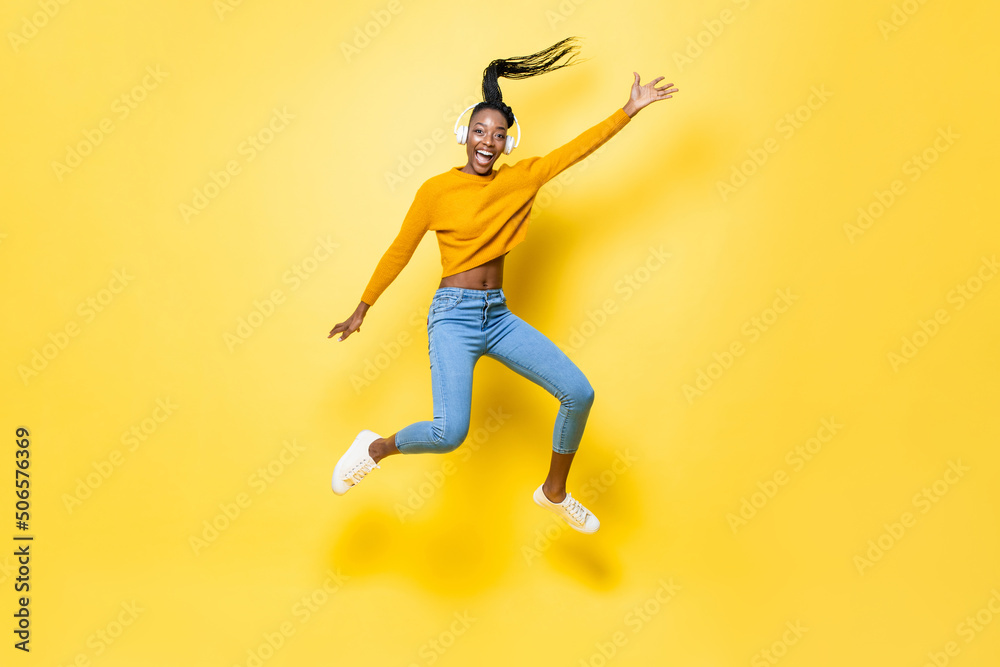 Positive enthusiastic young African American woman wearing headphones listening to music and jumping with hand up in yellow isolated studio background