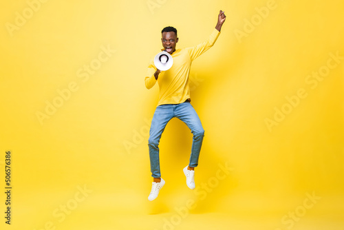 Powerful energetic young African man jumping and yelling on megaphone in studio yellow color isolated background