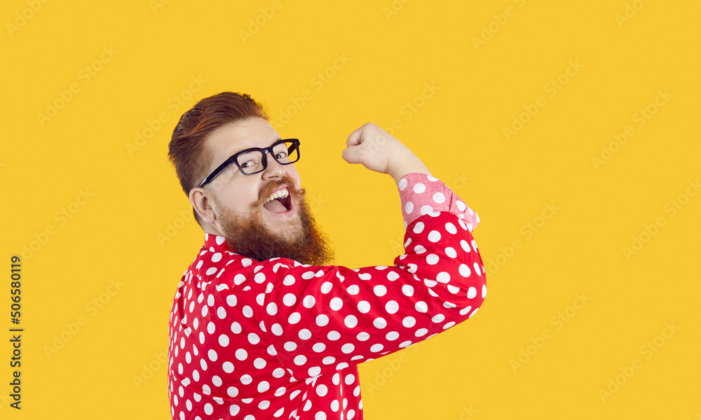 Funny stylish bearded chubby man with smile on face shows showing biceps on yellow background. Joyful crazy redhead millennial man in glasses and red shirt in white peas shows his power. Web banner. - obrazy, fototapety, plakaty 