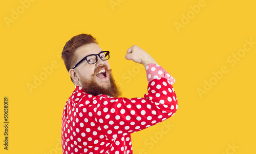 Funny stylish bearded chubby man with smile on face shows showing biceps on yellow background. Joyful crazy redhead millennial man in glasses and red shirt in white peas shows his power. Web banner. photo