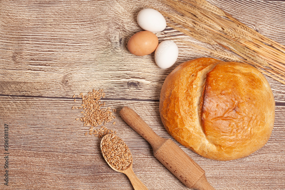 Loaf of round bread with egg, wheat ears, grains, rolling pin on a wooden table top view. Culinary background, baking concept Loaf of round bread with egg, wheat ears, grains, rolling pin on a wooden table top view. Culinary background, baking concept