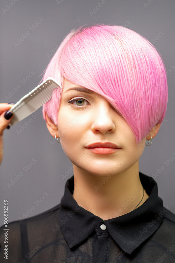 The female hairdresser is combing the dyed pink short hair of the young woman against the dark wall The female hairdresser is combing the dyed pink short hair of the young woman against the dark wall