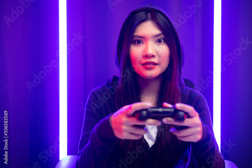 Playing video game. Young asian pretty woman sitting on chair holding joystick in living room. Happy female Professional Streamer chinese wearing hoodie playing game online in dark room neon light.