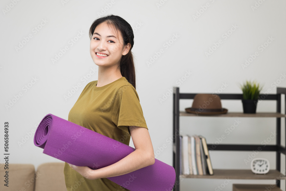 Young beautiful asian woman standing pose smiling to camera holding yoga mat. Cheerfully sporty female workout and exercise wearing sport wear. Charming woman relax after fitness training.