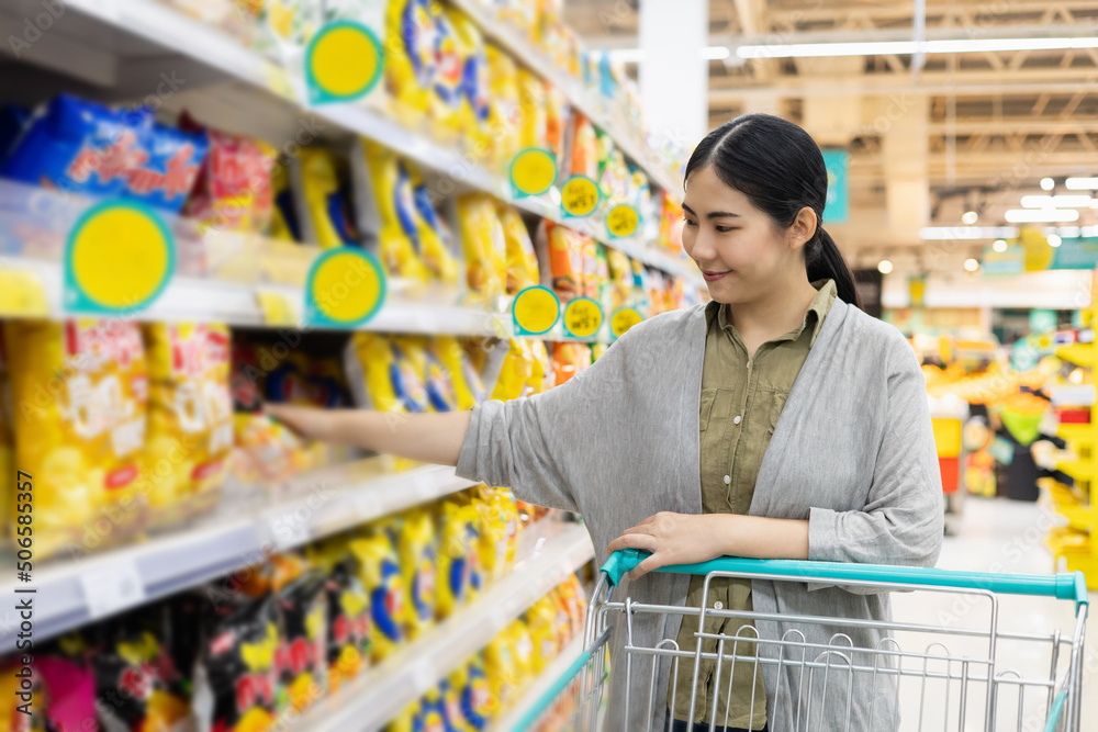 Asian woman shopping from the shelf to the shopping cart in supermarket modern trade hall. Female looking from a grocery store shelf and pick it up to trolley cart.