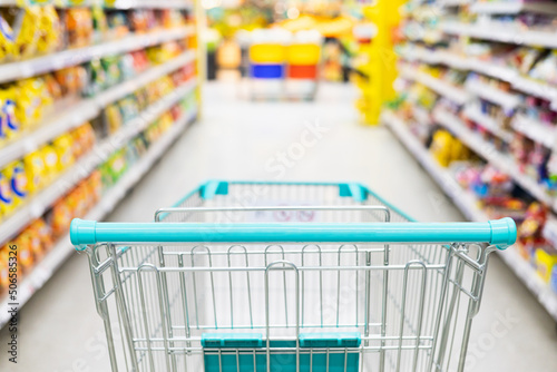 Supermarket aisle with empty green shopping cart. Trolley cart at the product shelf in the supermarket. Grocery store with many food and appliances in modern trade hall.