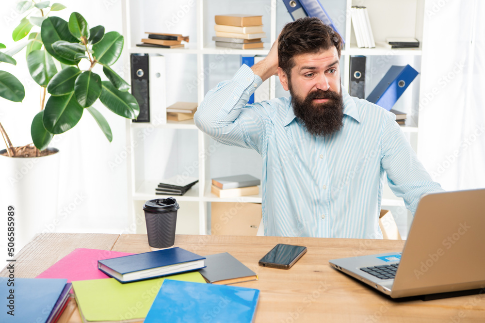 Confused businessman scratching head while working at laptop in office, confusion
