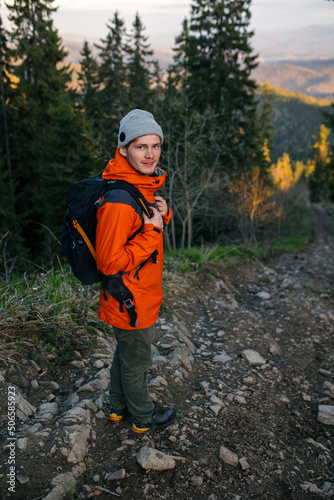 Young attractive guy in the mountains in an orange jacket and gray hat. Hiker with a backpack in the mountains at sunset.
