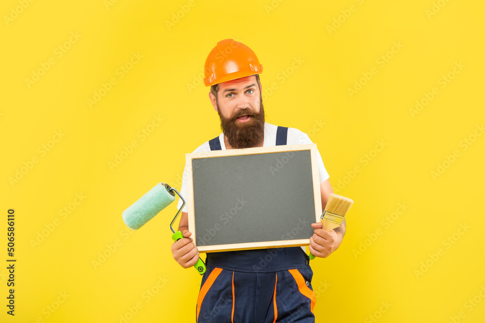 positive man in helmet and work clothes hold paint roller brush and blackboard with copy space positive man in helmet and work clothes hold paint roller brush and blackboard with copy space