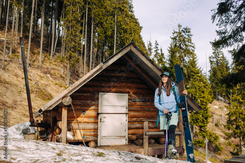 Attractive skier girl stands with skis in the spring near a forest house in the mountains.