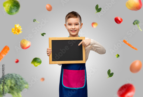 Fototapeta Naklejka Na Ścianę i Meble -  cooking, culinary and profession concept - happy smiling little boy in apron holding chalkboard over food on grey background