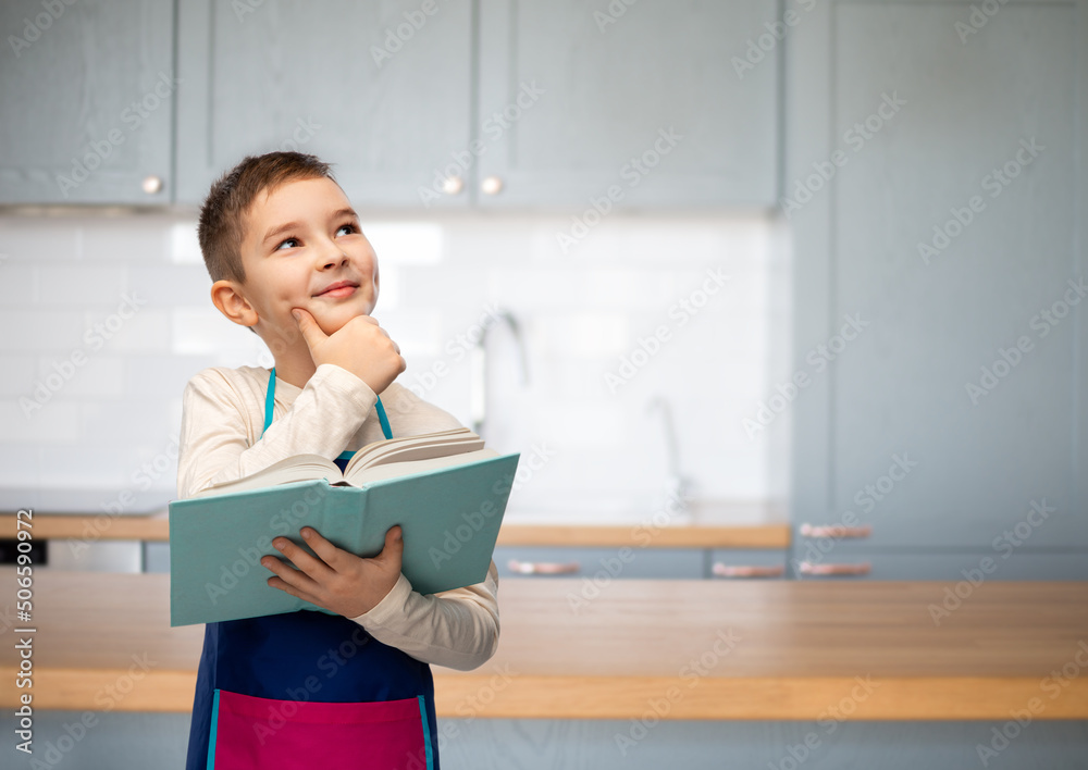 cooking, culinary and profession concept - happy smiling little boy in apron reading cook book and thinking over home kitchen background